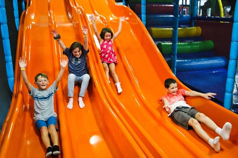Four kids racing down indoor slides side-by-side with their arms up, laughing — the kind of joyful active play Planet of Play is built around.